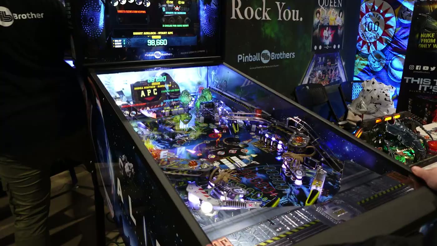 Máquina Alien de Pinball Brothers en pleno playfield, con el arte de Rock You de fondo