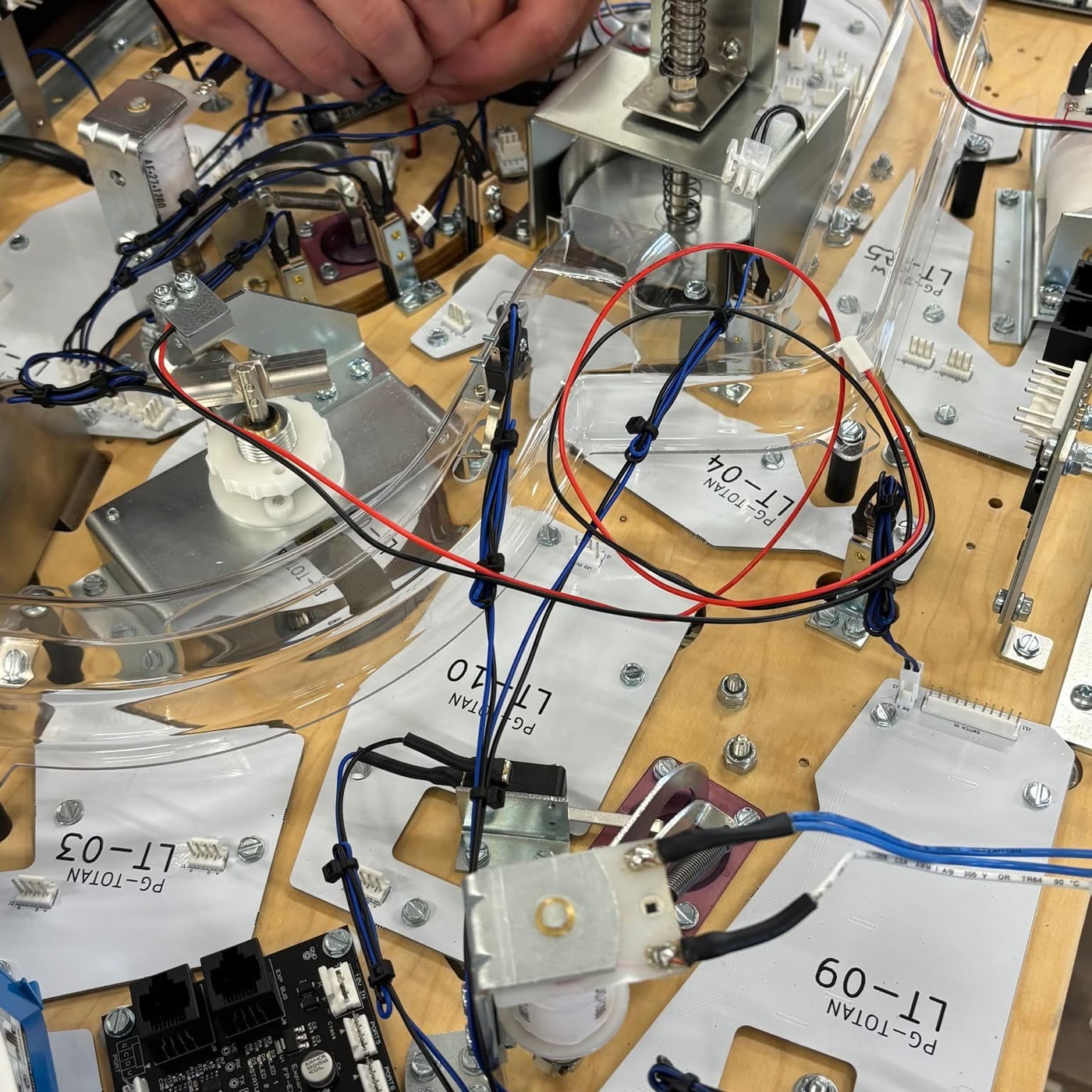 Manos de un técnico ajustando el cableado bajo el playfield, entre piezas etiquetadas como PG-TOTAN